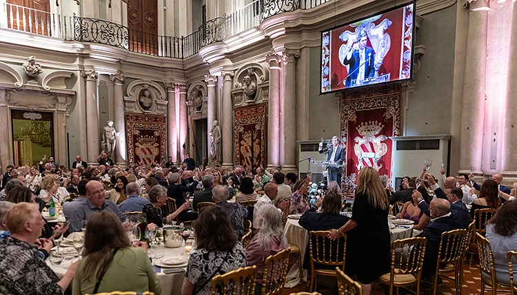 A large group of people are attending a formal dinner event in an ornate hall with a speaker at a podium and a large screen displaying the speaker’s image.