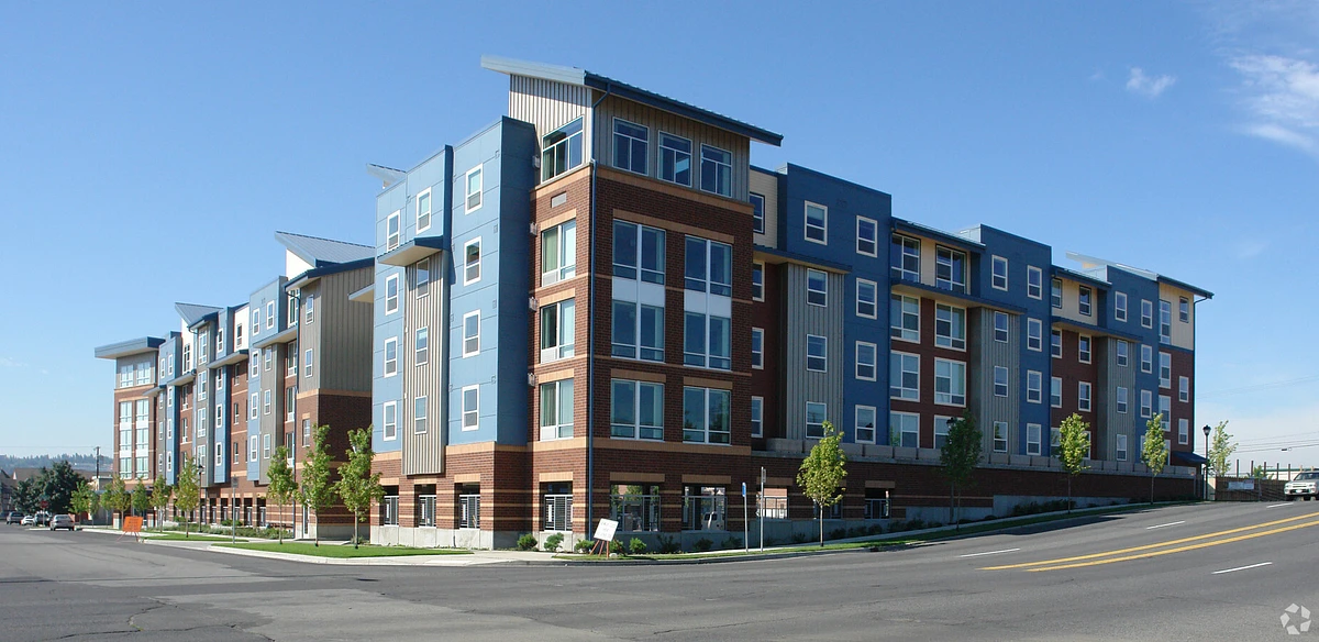 A modern, multi-story apartment building with a mixed-material facade sits alongside a sloping street under a clear blue sky.
