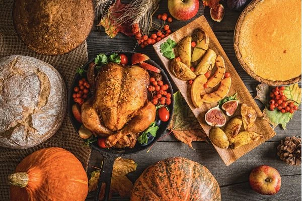 A festive autumn table spread featuring a roasted turkey, various breads, seasonal fruits, pumpkins, and a pumpkin pie.